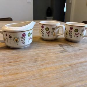 Op de afbeelding: Een crèmekleurige melkkan en twee bijpassende mokken. Elk stuk heeft een bloemenontwerp met roze, gele en groene bloemen. De mokken hebben een handvat en een bruine rand. De melkkan heeft een tuit en een handvat.
