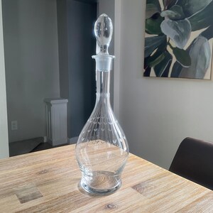 Puede incluir: Una jarra de vino de cristal transparente con tapón. La jarra tiene un cuello largo y un cuerpo bulboso con diseños de hojas grabadas. El tapón tiene forma redondeada. La jarra está sobre una mesa de madera.