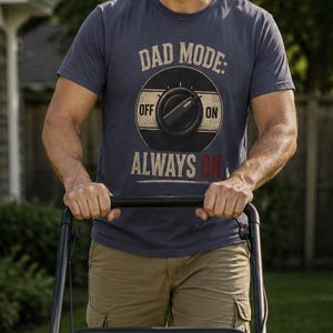 May include: A slate blue short-sleeve t-shirt featuring a graphic of a circular control knob with the text DAD MODE: OFF ON and ALWAYS ON printed in a vintage style. The design includes a black circle with a central dial and horizontal lines.