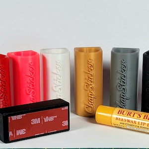 May include: Six different colored 3D printed lip balm holders with the text "Chap Sticker" on each. The holders are sitting on a white surface. A Burt's Bees Beeswax Lip Balm with Vitamin E & Peppermint is in the foreground.