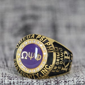 May include: A gold ring with a purple centre and the Greek letters Omega, Psi, and Phi. The ring is inscribed with "OMEGA PSI PHI FRATERNITY, INC." and "EN GRIMM CE DAWG". The ring is set with small, clear stones.