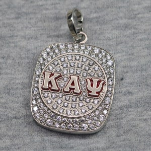 May include: A silver pendant with a red and white "ΚΑΨ" inscription surrounded by clear rhinestones.