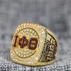 Könnte beinhalten: Ein goldfarbener Ring, besetzt mit kleinen, klaren Edelsteinen. Der Ring zeigt die Zahl "100" in Rot, umgeben von klaren Steinen. Die Seite des Rings hat Text in Dunkelbraun. Der Ring liegt auf einem grauen Stoff.