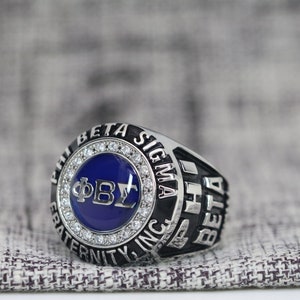 May include: A silver ring with a blue enamel center featuring the letters "ΦΒΣ" surrounded by a circle of small diamonds. The ring also has the words "Phi Beta Sigma Fraternity Inc." and "1934" engraved on the band.