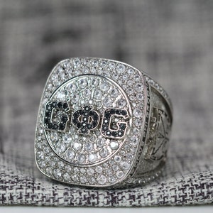 May include: A silver ring with a square face featuring a black and white 'G.O.G' inscription surrounded by clear rhinestones.
