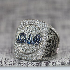 Może przedstawiać: Srebrny pierścień z dużym, kwadratowym frontem, na którym widnieją litery "Q.A.Φ" w niebiesko-białym szkliwie. Pierścień zdobią małe, przezroczyste kamienie wokół frontu i obrączki. Obrączka jest wygrawerowana słowami "MEGA LAMBDAZ", "INTEGRITY" i "BROTHERHOOD".