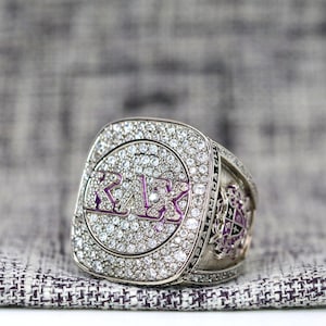 Può includere: Un anello da campionato in argento con un logo 'KSU' viola circondato da diamanti.