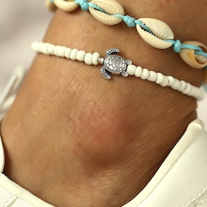 May include: White beaded ankle bracelet with a silver turtle charm. The bracelet is layered over a blue and white shell anklet.