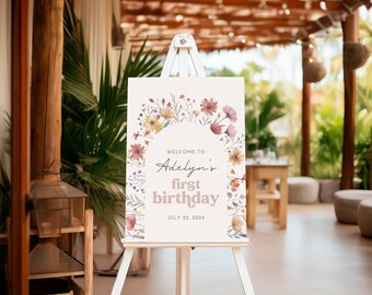 May include: A white sign with a floral border and the text "Welcome to Adelyn's first birthday July 22, 2024" on a white easel.