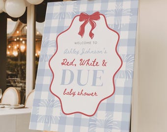 May include: A baby shower welcome sign on a wooden easel. The sign has a light blue gingham background with a red scalloped border. Text reads "Welcome to Ashley Johnson's Red, White & DUE baby shower."