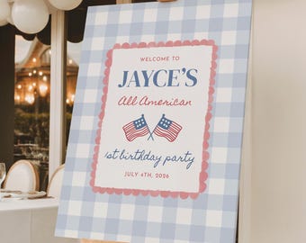 May include: A sign on a wooden easel with a blue and white gingham pattern. The sign reads "Welcome to Jayce's All American 1st birthday party July 4th, 2026" with two American flags. White balloons are above the sign.