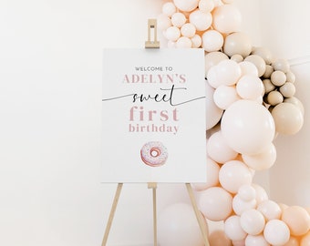 Könnte beinhalten: Willkommen zu Adelyns süßem ersten Geburtstagsschild mit einer rosafarbenen Donut-Illustration. Das Schild steht auf einem Holzstaffelei vor einer Wand, die mit einer Ballon-Girlande dekoriert ist.