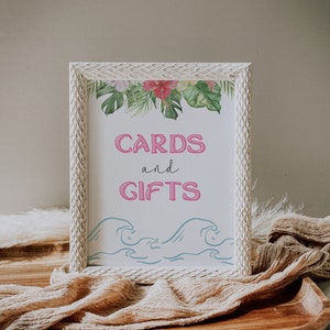 May include: White wooden frame with a sign that says "CARDS and GIFTS" in pink lettering. The sign is decorated with tropical leaves and flowers. There are blue waves at the bottom of the sign.