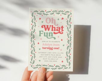 May include: A rectangular invitation card with a cream background, featuring a green leafy border with small pink flowers and red stars. The text reads: Oh What Fun, JOIN US TO CELEBRATE, Adelyn Jean turning one!, SATURDAY | DEC 10TH | 2-4 PM, 1234 NORTH POLE DRIVE, ANY CITY, ANY STATE 12345, RSVP TO EMILY AT (XXX) XXX-XXXX.