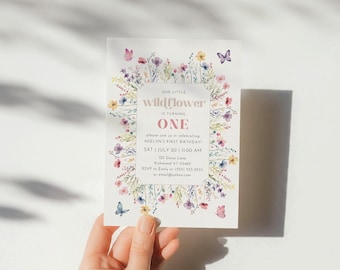 May include: A floral-themed first birthday invitation with a watercolor design. The invitation features a pink and white banner with the text "Our Little Wildflower is Turning One". The invitation also includes the date and time of the party, as well as the address and RSVP information.