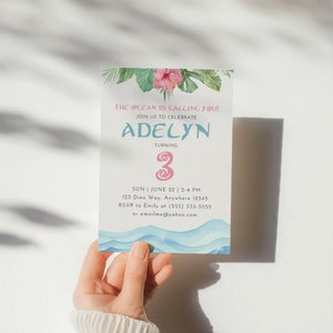 May include: A light blue and white invitation card with a watercolor design of ocean waves and tropical leaves. The text on the card reads "The ocean is calling you! Join us to celebrate Adelyn turning 3. Sun June 22 | 2-4 PM 123 Dino Way, Anywhere 12345 RSVP to Emily at (555) 555-5555 or emailme@yahoo.com"