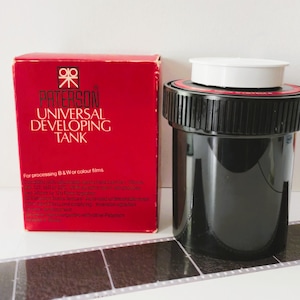 Könnte beinhalten: Ein schwarzer Paterson Universal Entwicklungstank mit weißem Deckel und einer roten Schachtel mit der Aufschrift Paterson Universal Developing Tank for processing B&W or colour films, begleitet von einem Streifen 35mm Film.