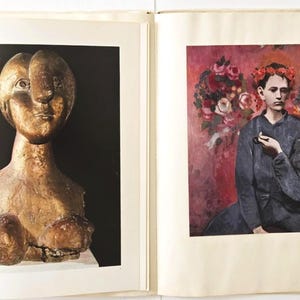 May include: An open book displaying two distinct art prints. The left page features a golden-toned, abstract bust sculpture against a black background. The right page shows a painting of a person wearing a red floral crown against a pink floral backdrop.