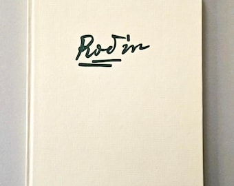 Op de afbeelding: Een hardcover boek met een crèmekleurige, textuurrijke stoffen kaft. Op de voorzijde staat het woord Rodin in een donkergroen, gestileerd cursief handschrift, met een horizontale lijn onder de tekst.
