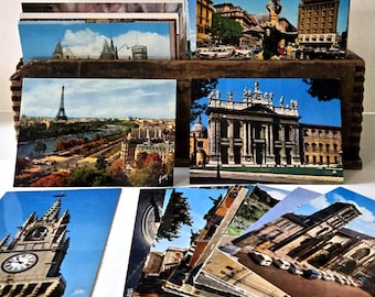 May include: A collection of vintage postcards featuring various European cityscapes. The postcards are stacked in a wooden container and fanned out, showcasing images of landmarks like the Eiffel Tower and ornate buildings.