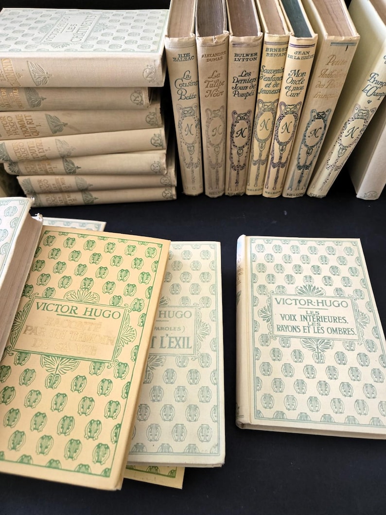 May include: A selection of vintage books with cream-coloured covers. The books, with French titles, display decorative patterns and text. Some are stacked, others upright, revealing their spines. The books are of various sizes.