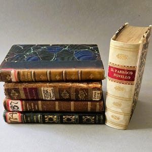 May include: A stack of four antique books with marbled covers and leather spines, positioned next to a single upright cream-colored book with a red label reading IL PARROCO NOVELLO, featuring gold embossed details and visible spine bands.