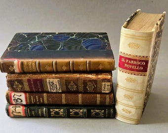 Op de afbeelding: Een stapel van vier antieke boeken met gemarmerde kaften en leren ruggen, naast een rechtopstaand crèmekleurig boek met een rood label met de tekst IL PARROCO NOVELLO, voorzien van gouden details en zichtbare ribben op de rug.