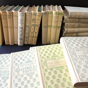 May include: A selection of antique books with cream-coloured covers and diverse titles. Some books are stacked, others stand upright. The covers display decorative patterns and text, including the author's name, Victor Hugo.