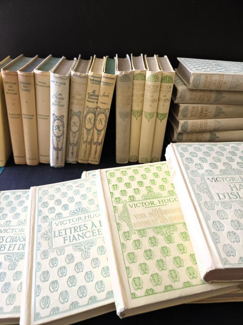 May include: A selection of antique books with cream-coloured covers and diverse titles. Some books are stacked, others stand upright. The covers display decorative patterns and text, including the author's name, Victor Hugo.