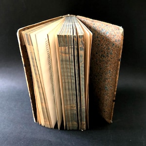 May include: An antique book with aged, yellowed pages and a worn, textured brown cover. The book is open, revealing the text and the edges of the pages. The cover has a mottled pattern with blue and brown tones.