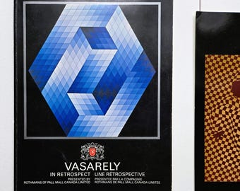 Op de afbeelding: Een zwarte boekomslag met de titel VASARELY IN RETROSPECT UNE RETROSPECTIVE PRESENTED BY ROTHMANS OF PALL MALL CANADA LIMITED. De omslag toont een groot blauw en wit geometrisch zeshoekig patroon bestaande uit kleine vierkantjes.