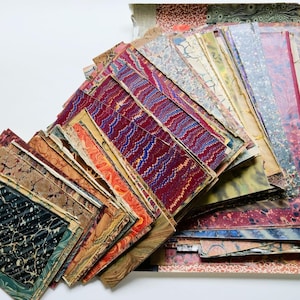 May include: A collection of rectangular paper sheets featuring various marbled patterns in colors including deep red, blue, black, orange, and cream. The sheets are stacked in an overlapping arrangement, displaying diverse swirling and wavy designs.