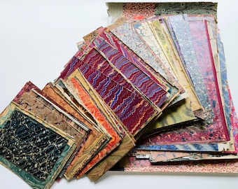 Op de afbeelding: Een verzameling rechthoekige vellen papier met diverse gemarmerde patronen in kleuren zoals dieprood, blauw, zwart, oranje en crème. De vellen liggen in een overlappende stapel en tonen uiteenlopende wervelende en golvende ontwerpen.