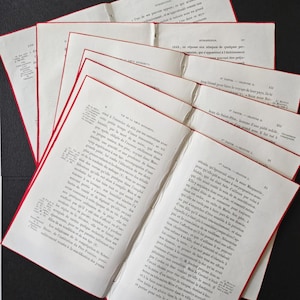 May include: Several open antique books with red-lined pages are fanned out on a dark surface. The pages are filled with small text, suggesting a historical or literary theme. The books appear aged.