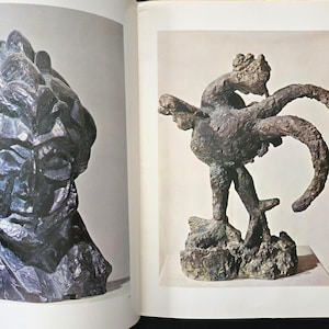 May include: An open book page displaying two sculptures. On the left, a dark, textured bust with angular features. On the right, a bronze-toned sculpture of a rooster with a rough, uneven surface standing on a base. The text "COLOR PRINTS" is visible.