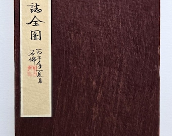Op de afbeelding: Een rechthoekig, donkerbruin boek met een houtnerftextuur. Een verticaal beige label aan de linkerkant bevat Japanse tekst in zwarte kalligrafie en rode stempels. De kaft van het boek is effen, rijk bruin.