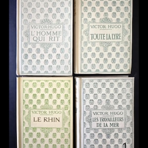 May include: Four vintage books by Victor Hugo. Each book has a cream-coloured cover with a repeating pattern of small, stylised floral designs. The titles include "L'Homme Qui Rit", "Toute La Lyre", "Le Rhin", and "Les Travailleurs De La Mer".