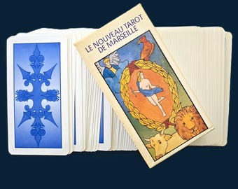 Op de afbeelding: Een tarotkaartspel met een blauw en wit ontwerp op de achterkant van de kaarten. De kaartdoos leest "LE NOUVEAU TAROT DE MARSEILLE" en bevat illustraties van figuren en symbolen. Het spel wordt gepresenteerd op een donkerblauwe achtergrond.