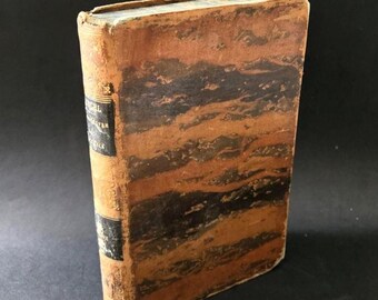 Op de afbeelding: Antiek boek met een versleten, getextureerde leren kaft. De kaft is voornamelijk bruin met donkerbruine en zwarte horizontale banden. De rug heeft een titel etiket en zwarte banden. De randen van het boek vertonen tekenen van slijtage.