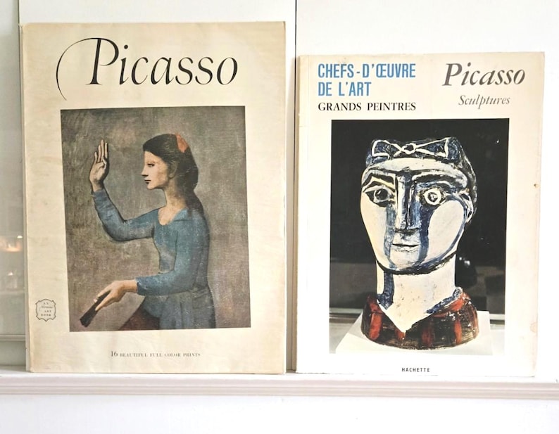 May include: Two softcover books featuring Picasso art. The left book cover shows a painting of a person in a blue shirt with the text Picasso and 16 beautiful full color prints. The right book cover displays a sculpture with the text Chefs-d'oeuvre de l'art Grands Peintres Picasso Sculptures Hachette.