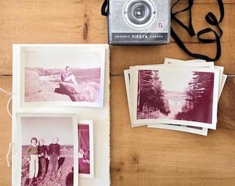 Op de afbeelding: Een vintage Kodak Brownie Fiesta camera met een zwarte band ligt op een houten oppervlak naast een stapel foto's en een fotoalbum. De foto's zijn in roze en witte tinten.