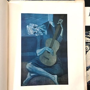 May include: A printed reproduction of Pablo Picasso's The Old Guitarist, featuring a monochromatic blue-toned painting of a seated, hunched figure playing a guitar. The artwork is centered on a white page within an open book.