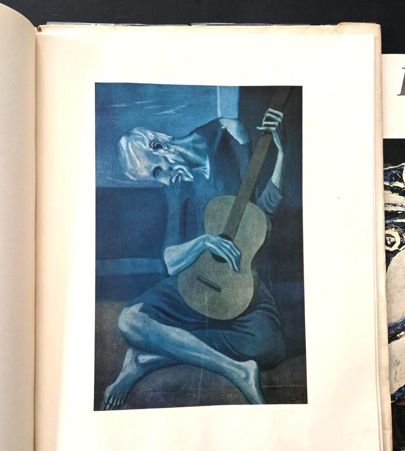 May include: A printed reproduction of Pablo Picasso's The Old Guitarist, featuring a monochromatic blue-toned painting of a seated, hunched figure playing a guitar. The artwork is centered on a white page within an open book.