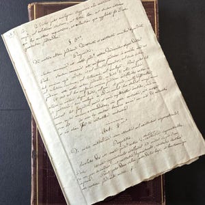 May include: An antique handwritten document with faded ink, placed on top of a dark brown leather-bound book. The paper is yellowed with age, and the script is in a cursive style. The document appears to be a legal or historical text.