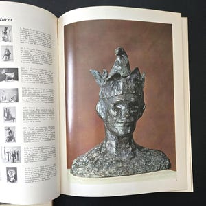 May include: A bronze-toned bust sculpture of a person wearing a pointed, multi-pointed hat with a textured, uneven surface. The sculpture is displayed on a page within an open book, which also features a list of other artworks.