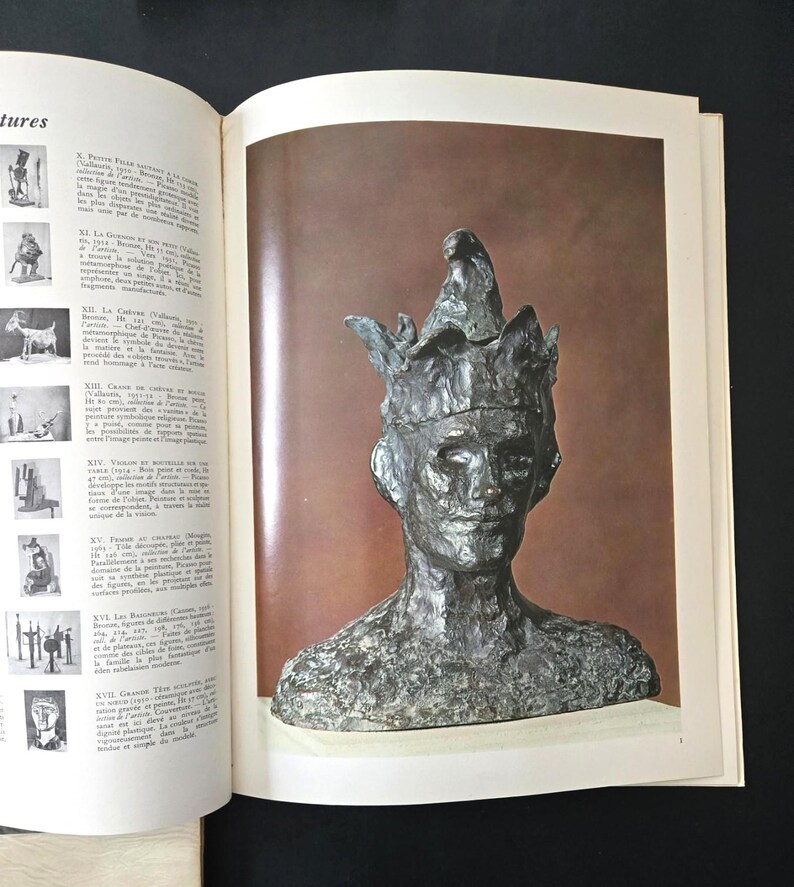 May include: A bronze-toned bust sculpture of a person wearing a pointed, multi-pointed hat with a textured, uneven surface. The sculpture is displayed on a page within an open book, which also features a list of other artworks.