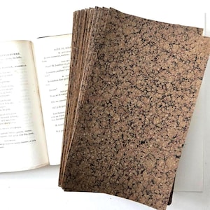 May include: A stack of rectangular paper sheets featuring a brown and black marbled pattern with intricate, vein-like details. Beside the stack lies an open book with visible printed text in French, including the heading Acte IV, Scene.