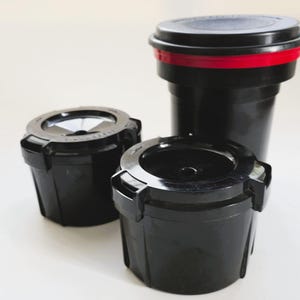 Op de afbeelding: Drie zwarte plastic filmontwikkelingstanks voor analoge fotografie. Twee kleinere tanks staan op de voorgrond met ronde deksels, terwijl een hogere tank met een rode ring rond het deksel erachter is geplaatst. Alle items zijn cilindrisch.