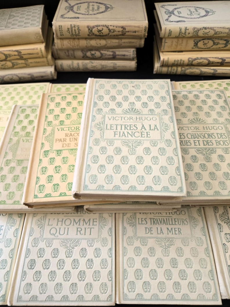 May include: A selection of vintage French books by Victor Hugo. The books feature off-white covers with embossed patterns and titles in French, including "Lettres &agrave; la Fianc&eacute;e". The books are stacked and arranged on a dark surface, evoking a classic literary aesthetic.