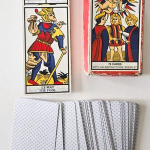 May include: A deck of Tarot cards with a red and white box labeled "L'ART TAROT OF MARSEILLES". The cards feature illustrations and text, including "LE MAT THE FOOL". A stack of cards is visible, along with a deck of cards with a gray pattern.